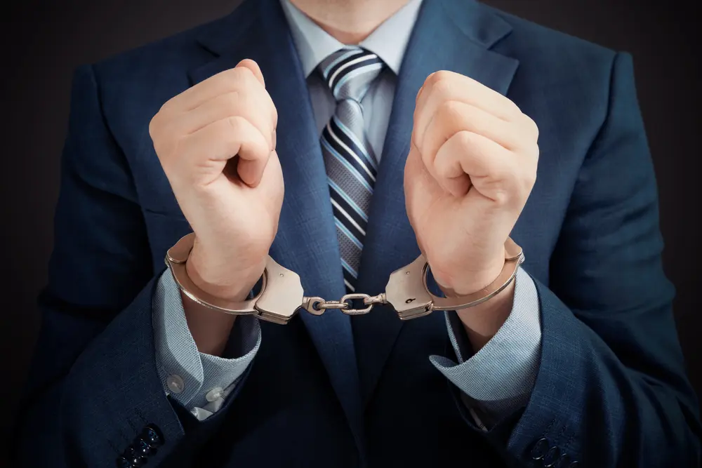 Businessman in a suit with handcuffs on his wrists, symbolizing arrest for corruption or white-collar crime.