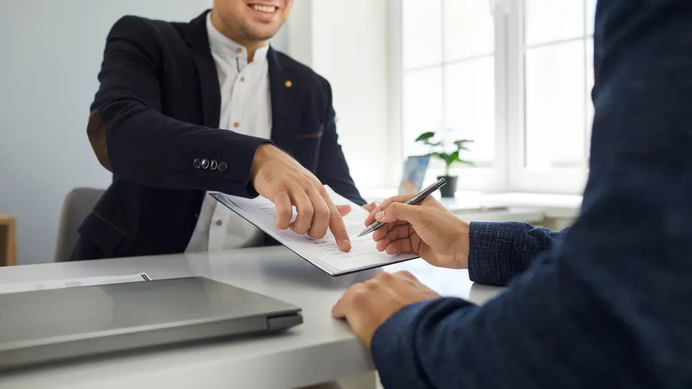 Smiling business agent guiding a happy client to sign a contract at an office desk. Smiling business agent guiding a happy client to sign a contract at an office desk.