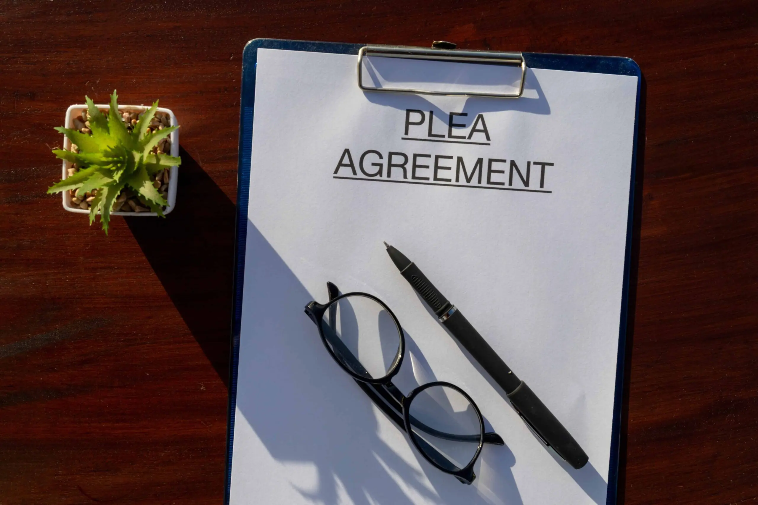 Plea agreement document on clipboard with pen and glasses on desk.