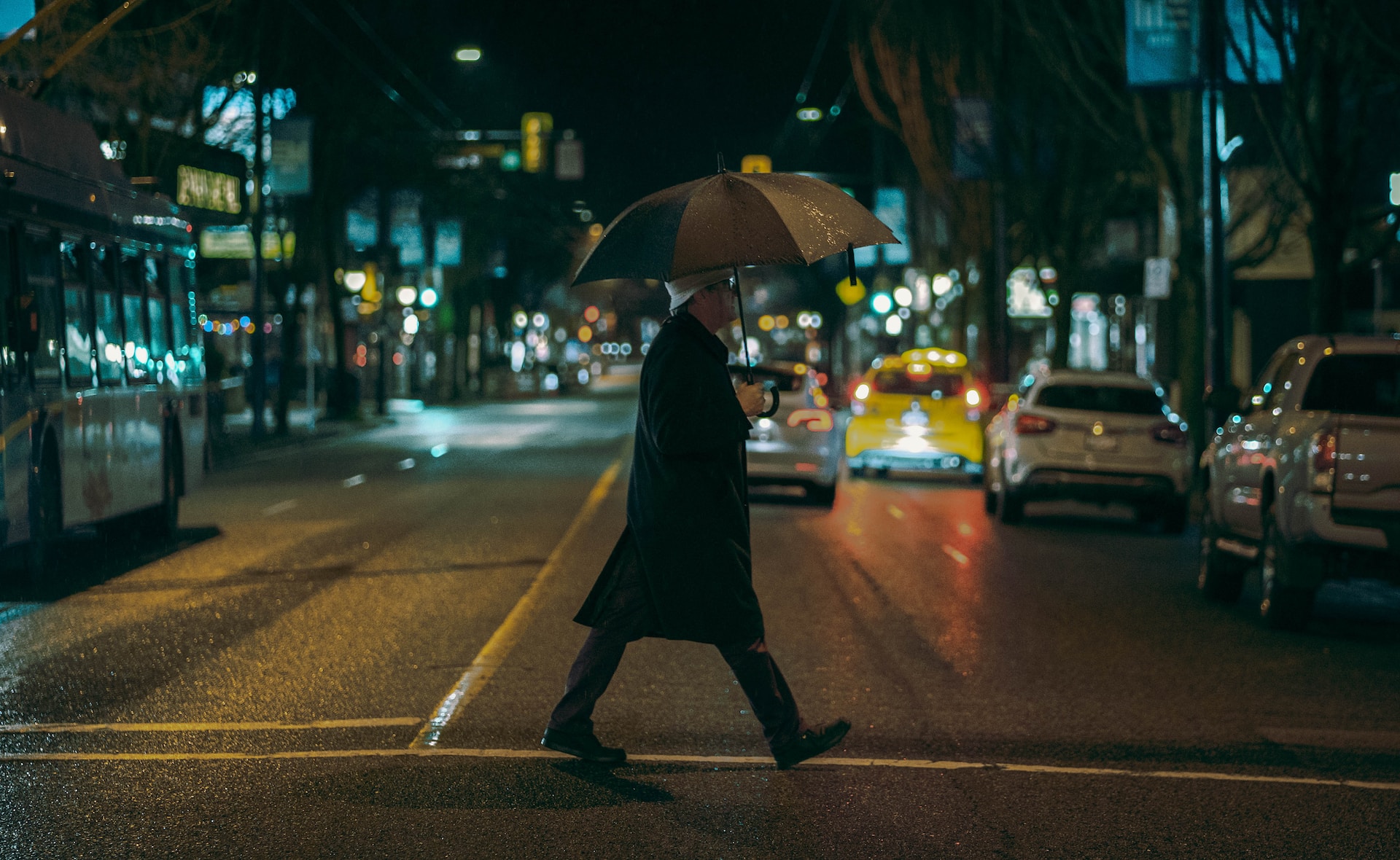 Person in black coat holding an umbrella and crossing the street at night